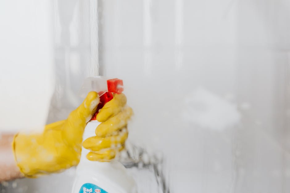 Close-up of a person wearing yellow rubber gloves holding a white spray bottle with a red nozzle, actively cleaning a glass shower door with a spray of cleaning solution visible on the surface. The ba