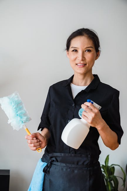 Close-up of a person wearing a denim shirt and pink cleaning gloves, holding a blue spray bottle of cleaning solution in their right hand. The background features a modern living room with a dark gray