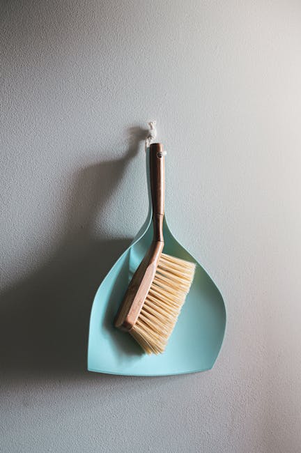 A light blue dustpan with a wooden handle and a matching brush with cream bristles hang on a white wall, illuminated by soft natural light, suggesting a clean and well-maintained domestic cleaning env