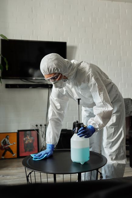 A professional cleaner wearing a white protective suit, orange gloves, and white sneakers is operating a yellow and gray vacuum cleaner on a tiled living room floor. The room features large glass slid